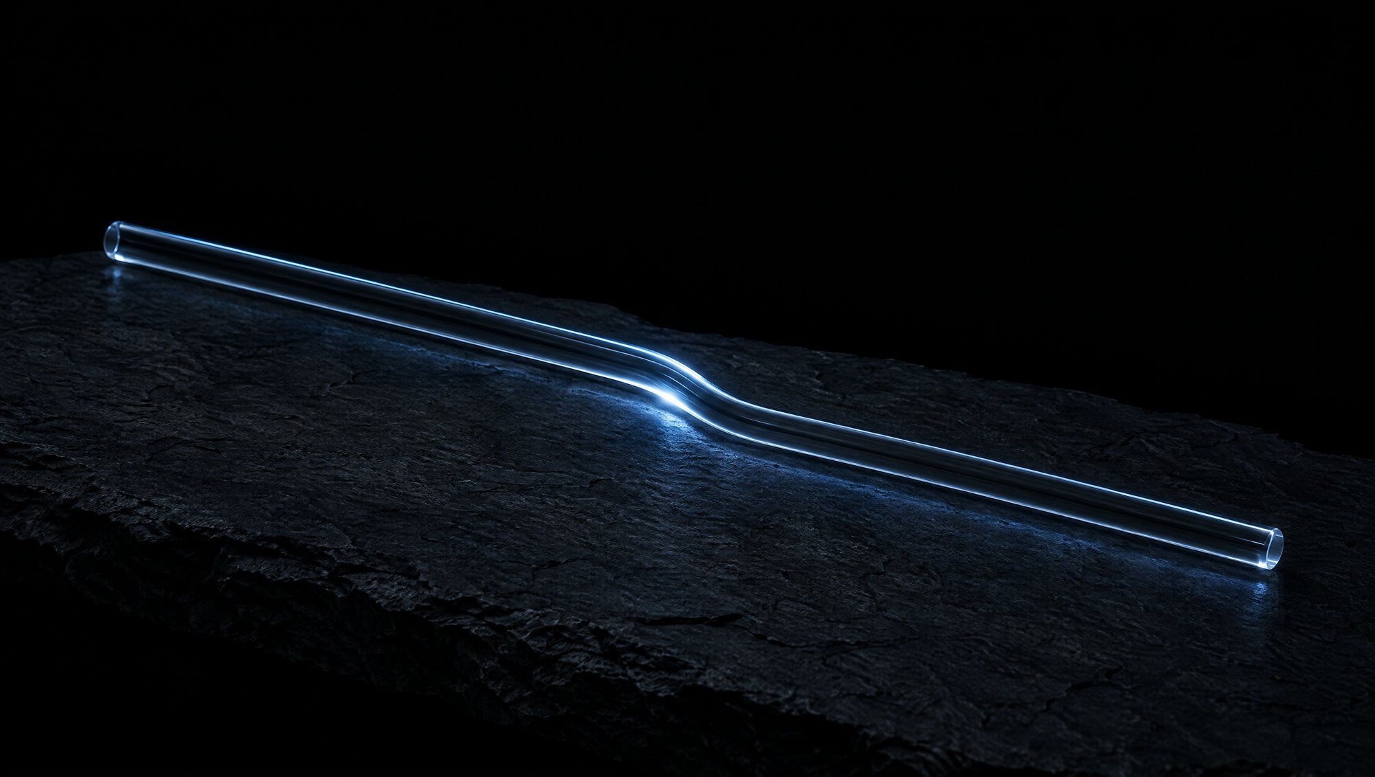 A long, slender clear glass tube with a gentle bend in the middle, lying diagonally across rough dark slate against black, its edges and bend lit by cool slate-blue light.