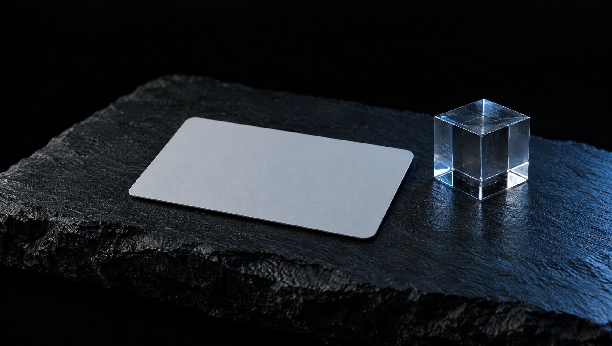 A blank pale grey card with rounded corners lying on rough dark slate beside a small clear glass cube, lit from the right by cool slate-blue light against a black background.