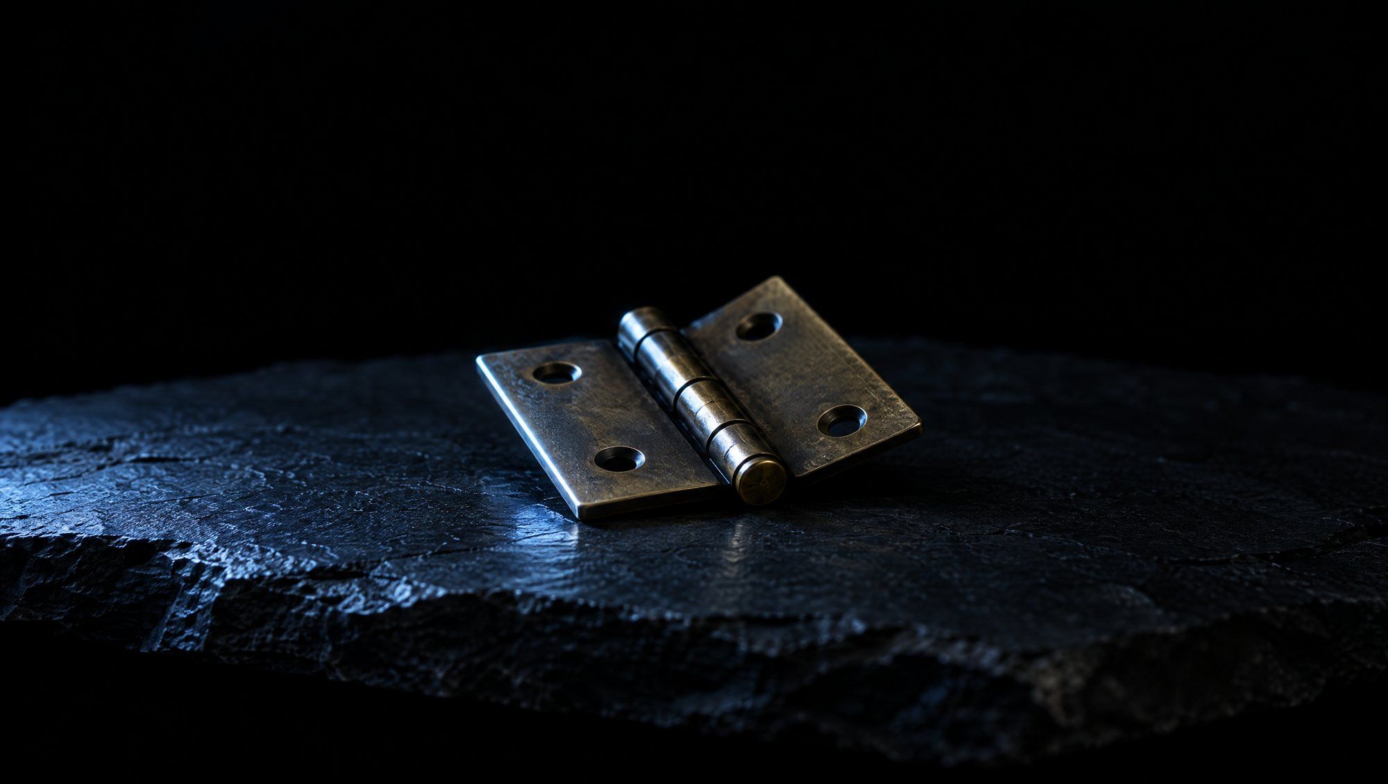 A small brass hinge lying half open on a rough slab of dark slate against black, its leaves and pin lit by cool slate-blue light from the left.