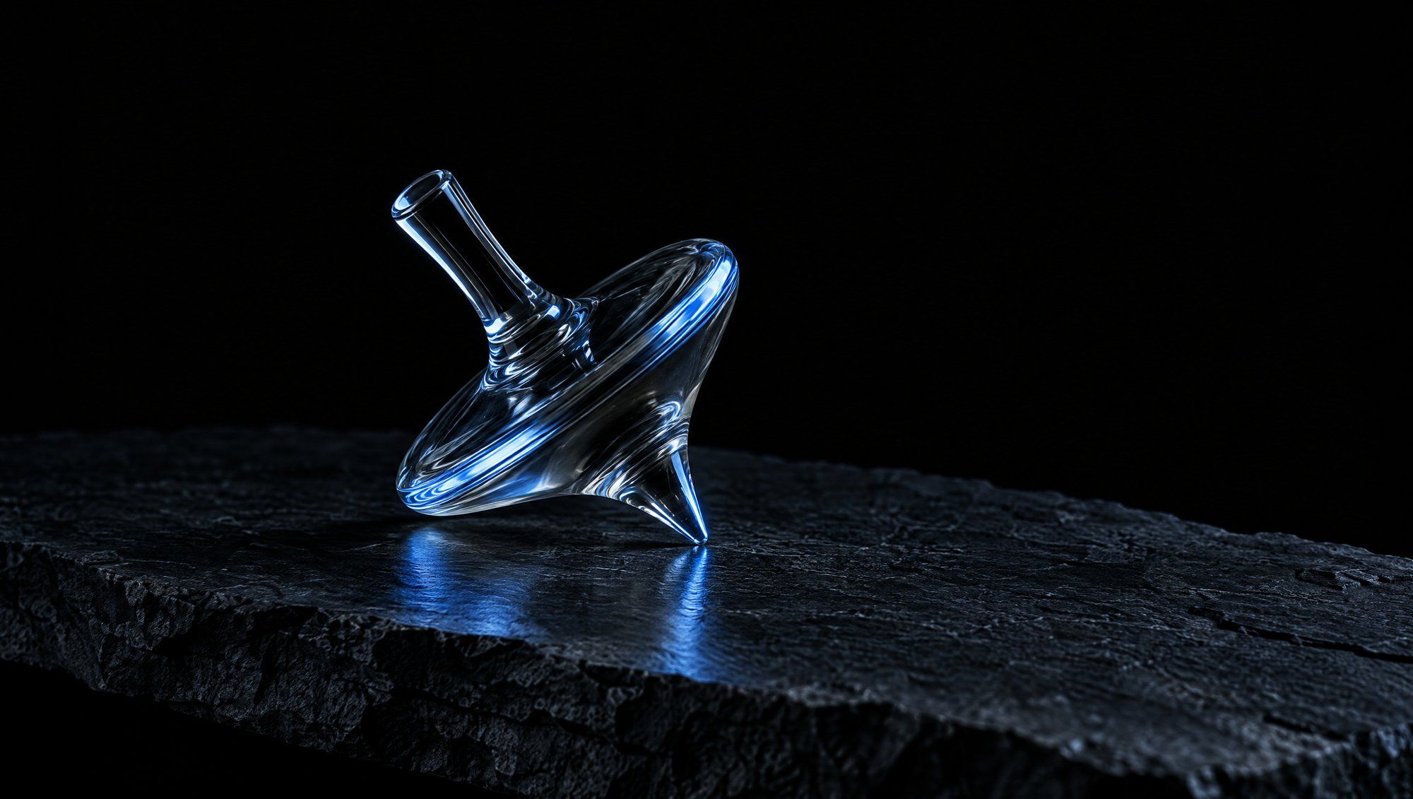 A clear glass spinning top tilted at an angle and resting on its point on a rough dark slate slab against black, its rim and point lit by cool slate-blue light that reflects on the stone.
