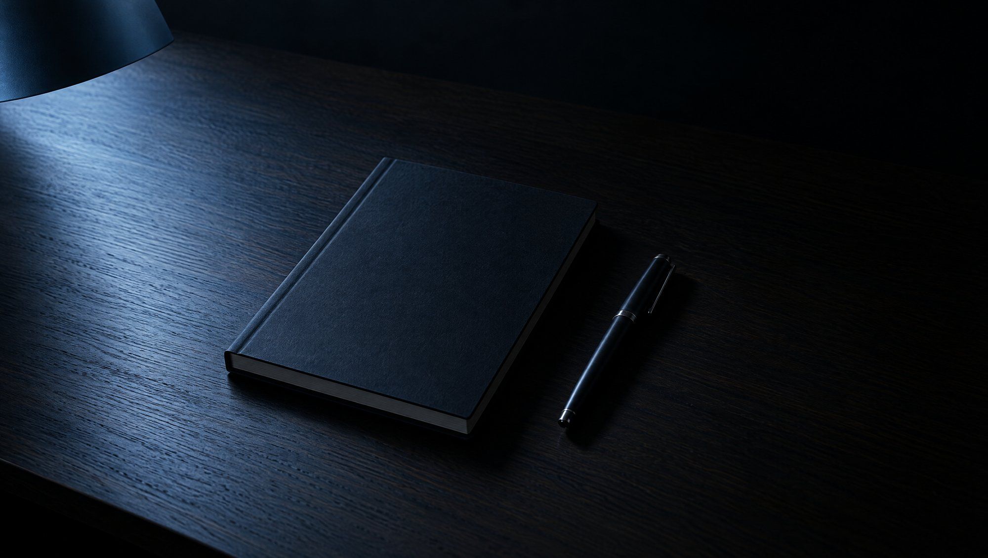 A closed dark notebook and a black pen on a dark wooden desk under a lamp's cool blue light.