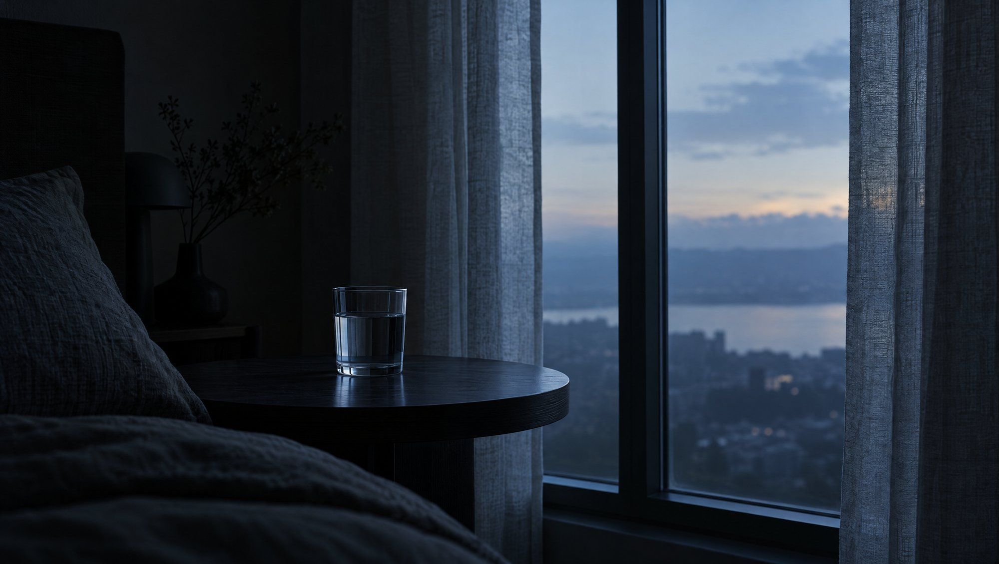 A plain glass of water on a bedside table by a tall window at dawn, cool slate-blue light falling through linen curtains into a dark room.