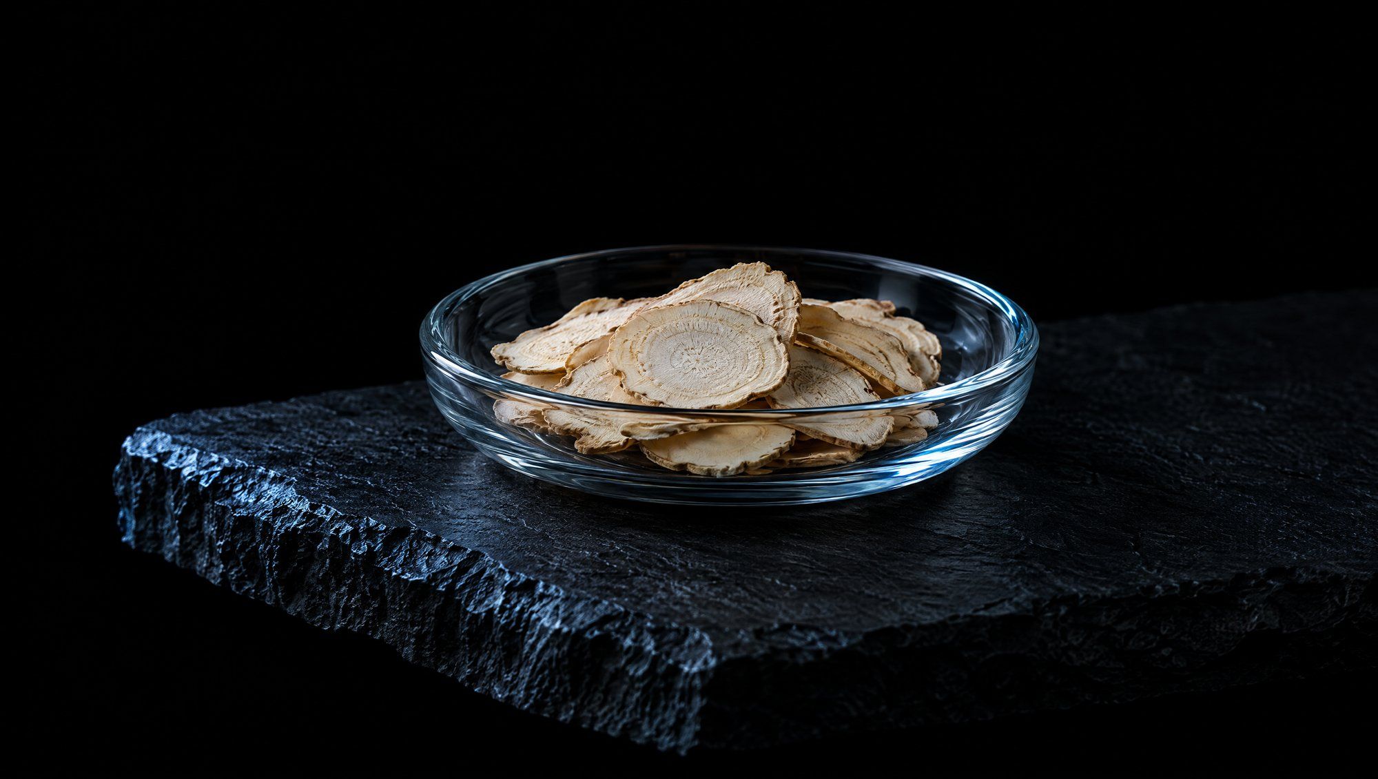 A shallow clear glass dish of thin, pale dried ginseng root slices on a rough dark slate slab against a black background, the glass rim and stone edge lit by a cool slate-blue light.