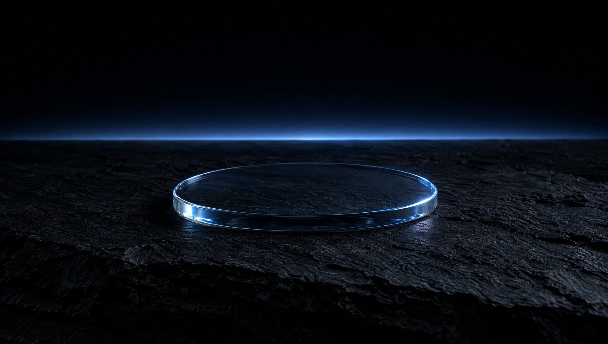 A flat clear glass disc lying on rough dark slate, with a thin band of cool slate-blue light along the horizon behind it against a black sky.