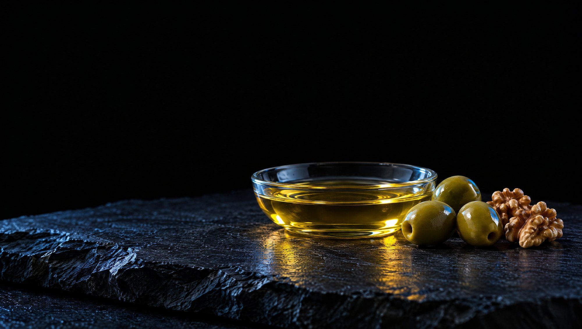 A shallow clear glass dish of golden olive oil beside three green olives and two walnut halves on rough dark slate against black, lit from the side with a cool slate-blue edge light on the stone.