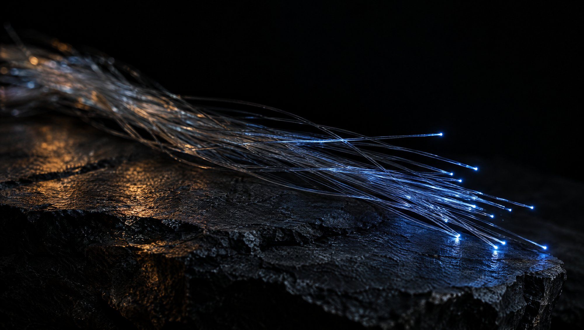 A loose bundle of fine clear glass fiber strands lying across rough dark slate against black, a few of them glowing slate-blue at their tips, with a faint amber reflection on the stone.
