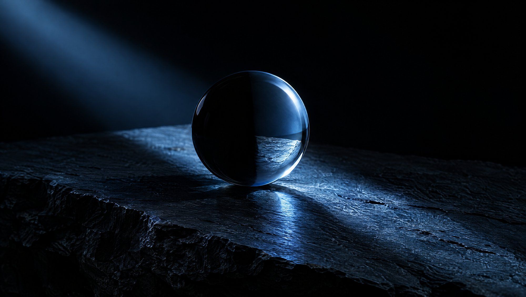 A single clear glass sphere on rough dark slate against black, one half in shadow and the other half lit by a diagonal beam of cool slate-blue light that falls across the stone.