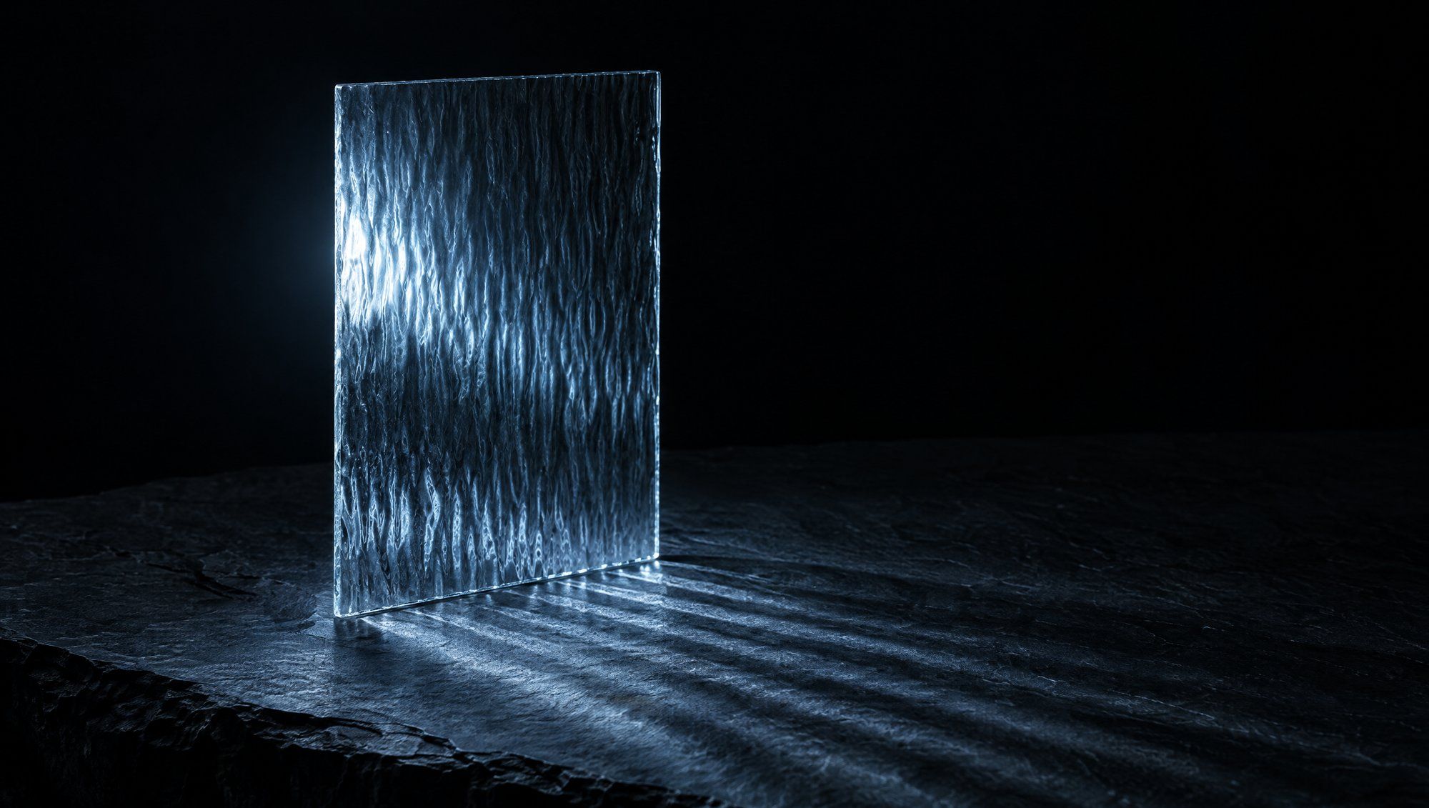 A pane of rippled glass standing upright on dark slate against black, with cool slate-blue light passing through it and falling across the stone in soft bands.