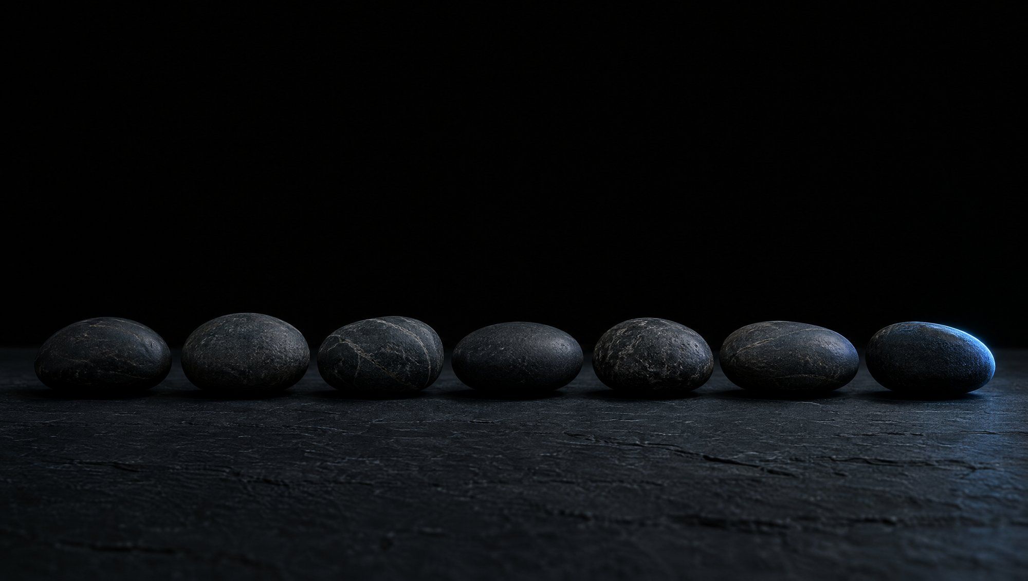 A row of seven smooth dark river stones on a slate surface, only the last stone touched by cool slate-blue light.