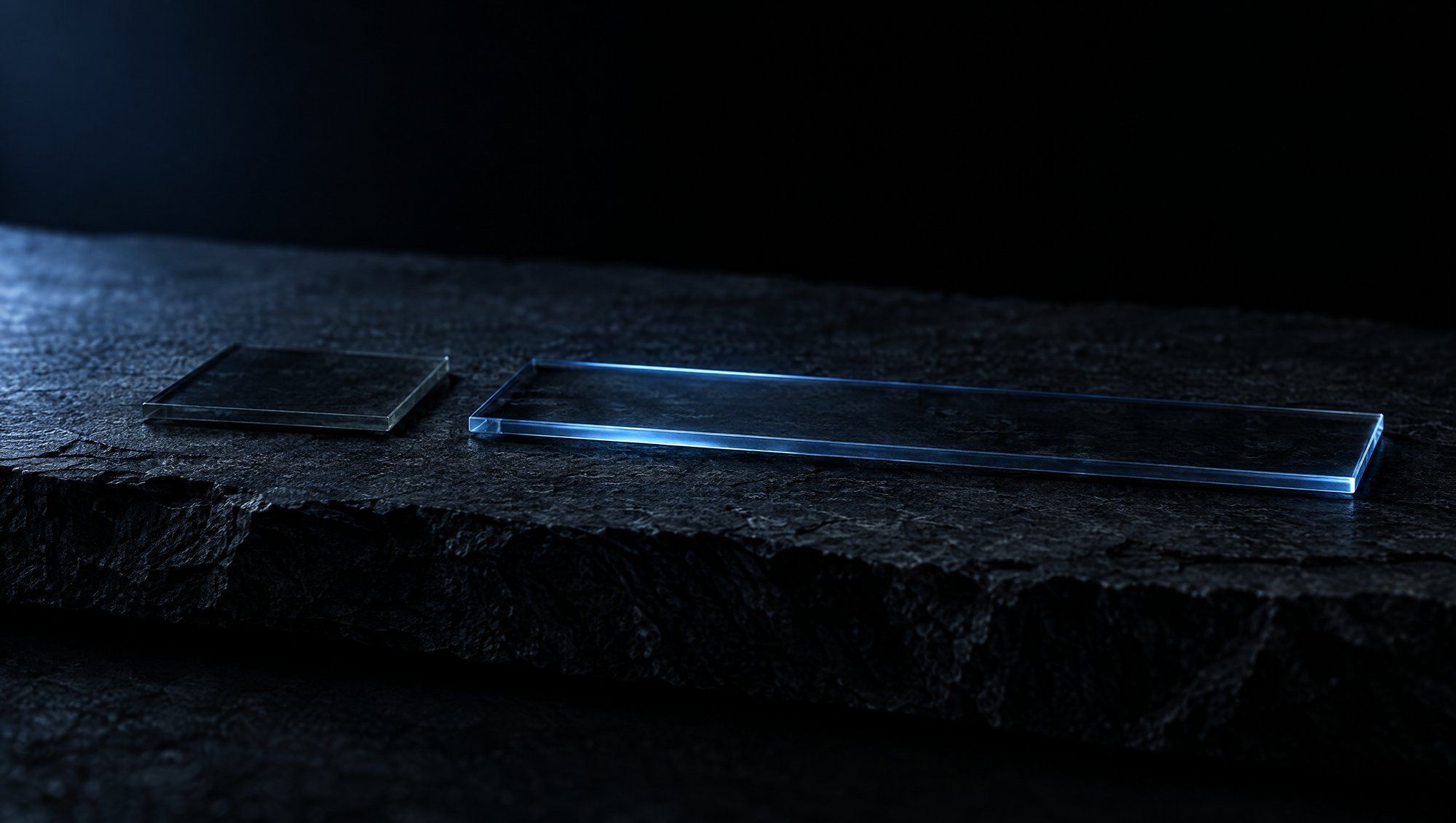 Two thin clear glass slabs lying flat on rough dark slate against black, a short square one on the left in shadow and a long narrow one on the right with its edges lit in cool slate-blue light.