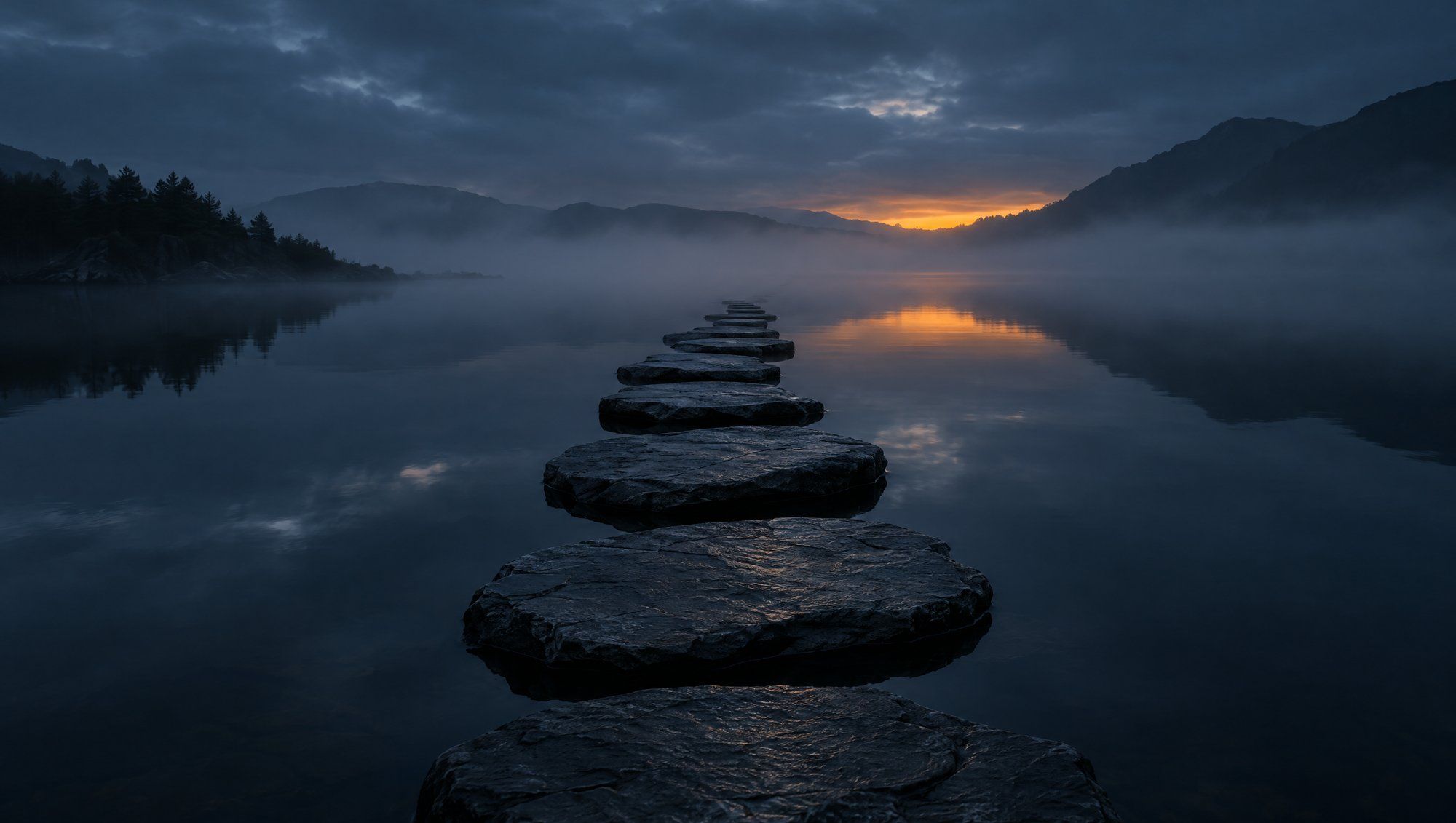 A line of flat dark stepping stones crossing still water at dusk toward misty hills, the sky slate blue with a faint amber glow on the horizon.