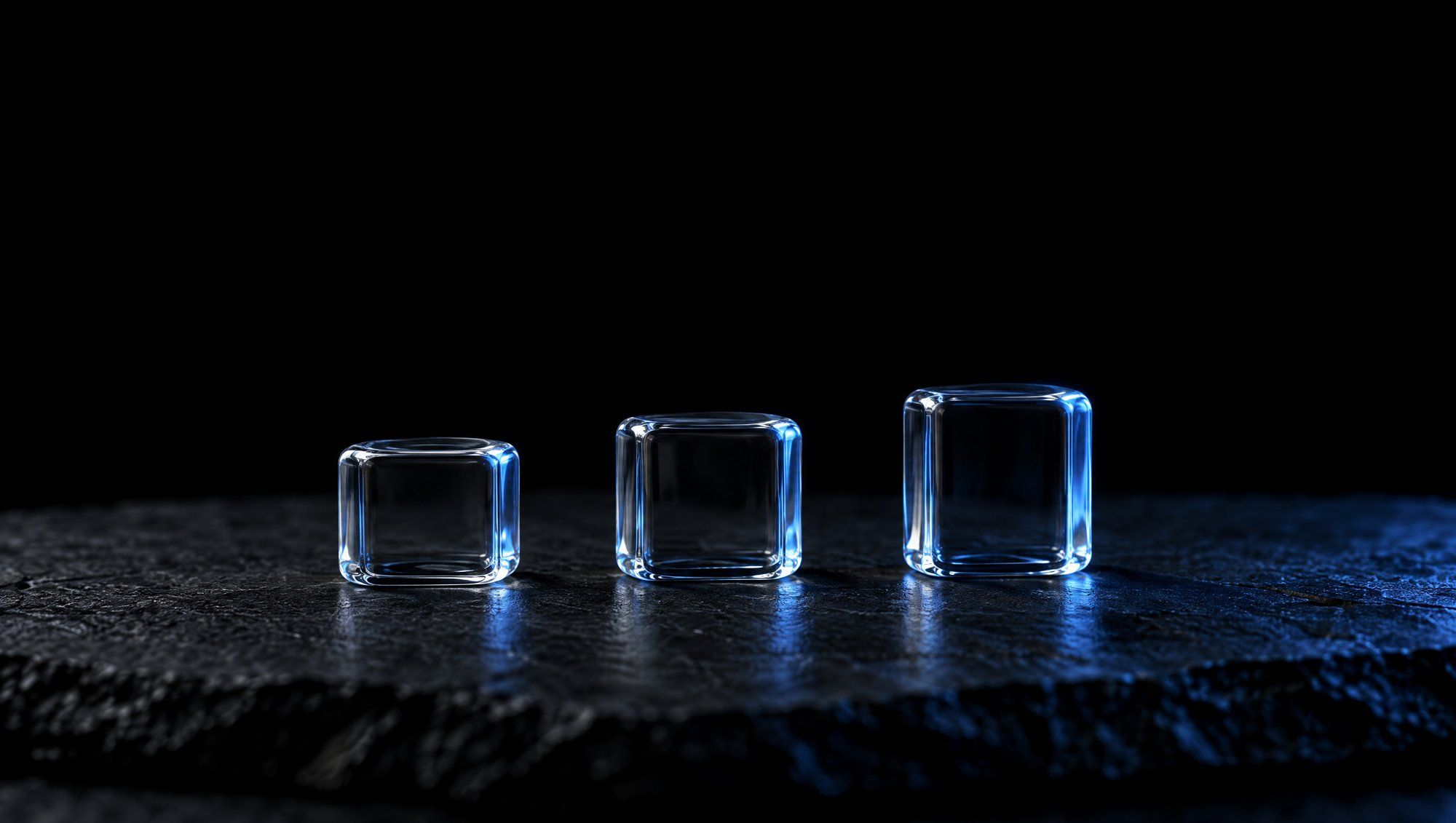 Three small clear glass cubes of slightly different heights in a row on rough dark slate against black, their edges lit by a cool slate-blue light.