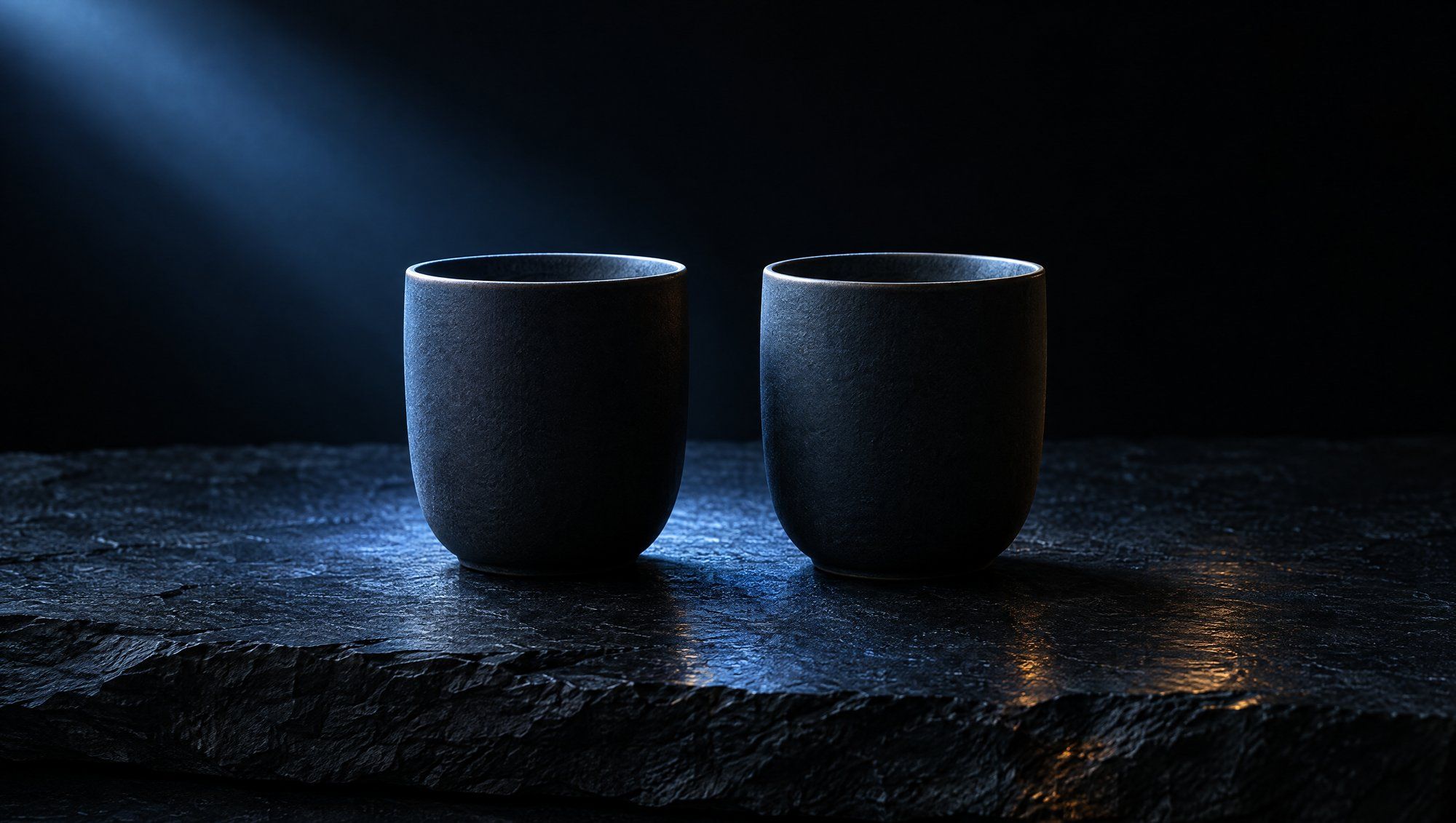 Two dark matte ceramic cups without handles standing side by side on rough dark slate against black, a beam of cool slate-blue light from the upper left catching their rims, with a faint amber reflection on the stone.