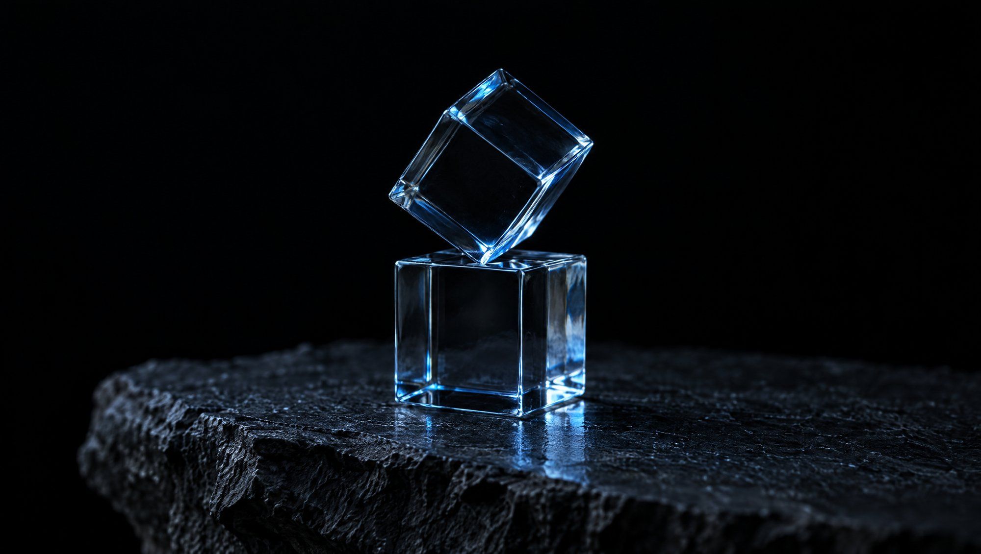 Two small clear glass cubes on rough dark slate against black, the top cube tilted and balanced on one edge of the lower cube, their edges lit with cool slate-blue light.