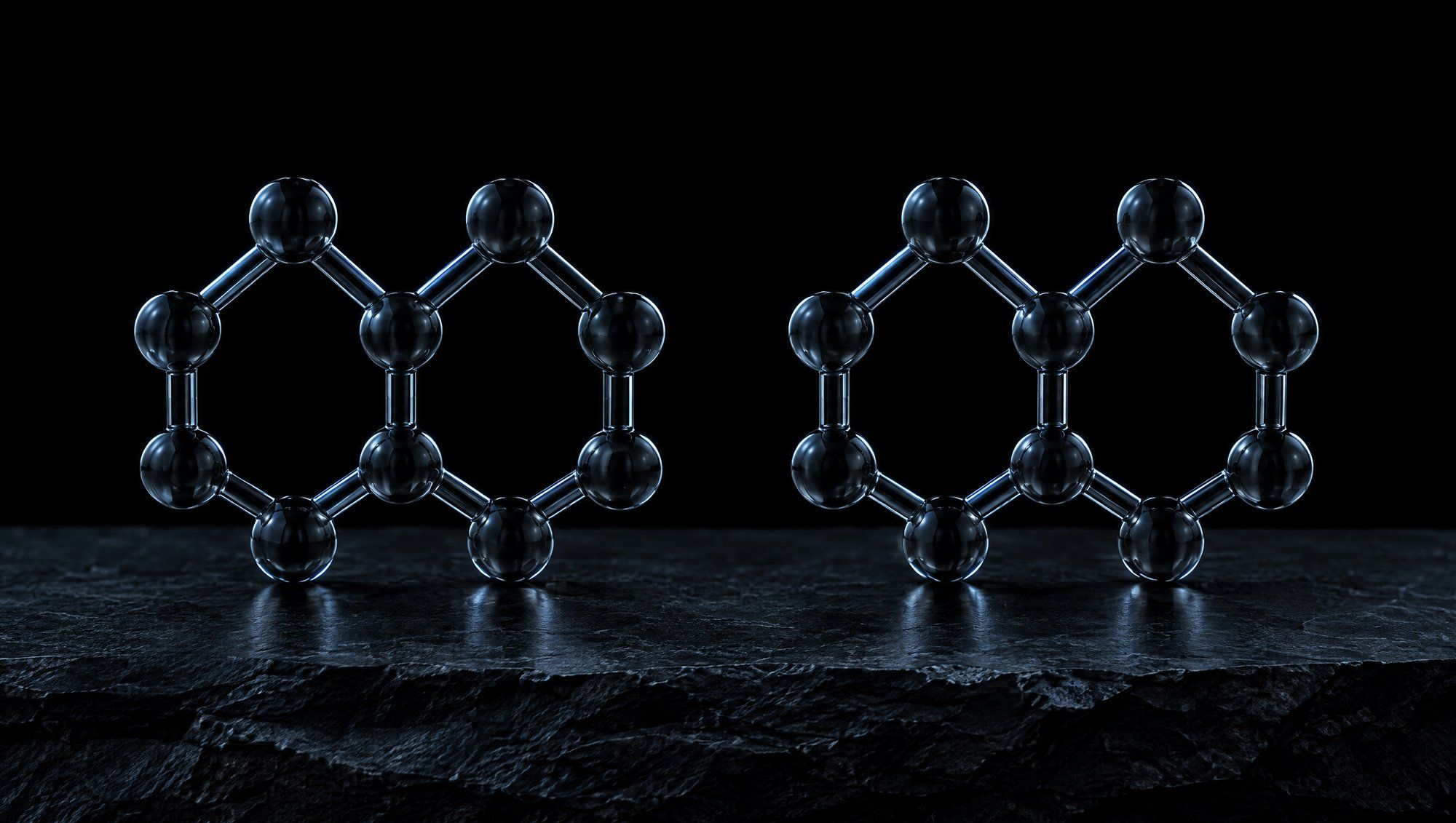 Two identical smoky glass molecular models of joined hexagonal rings standing side by side on dark slate stone against black, their edges lit in cool slate-blue light.