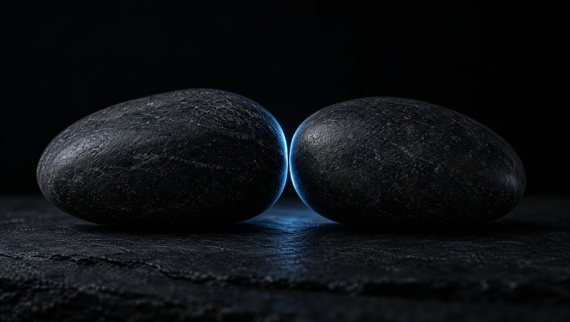 Two smooth dark river stones almost touching on a dark slate surface, a thin line of cool slate-blue light glowing in the narrow gap between them.